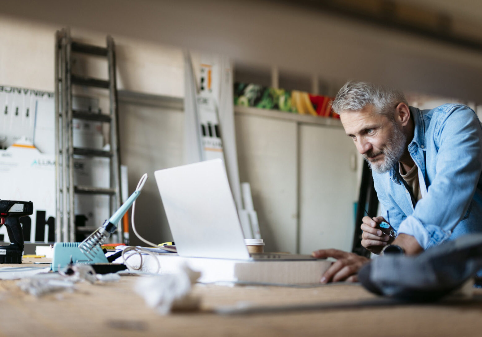 Mature craftsman concentrating on his laptop in a busy workshop, surrounded by tools and materials